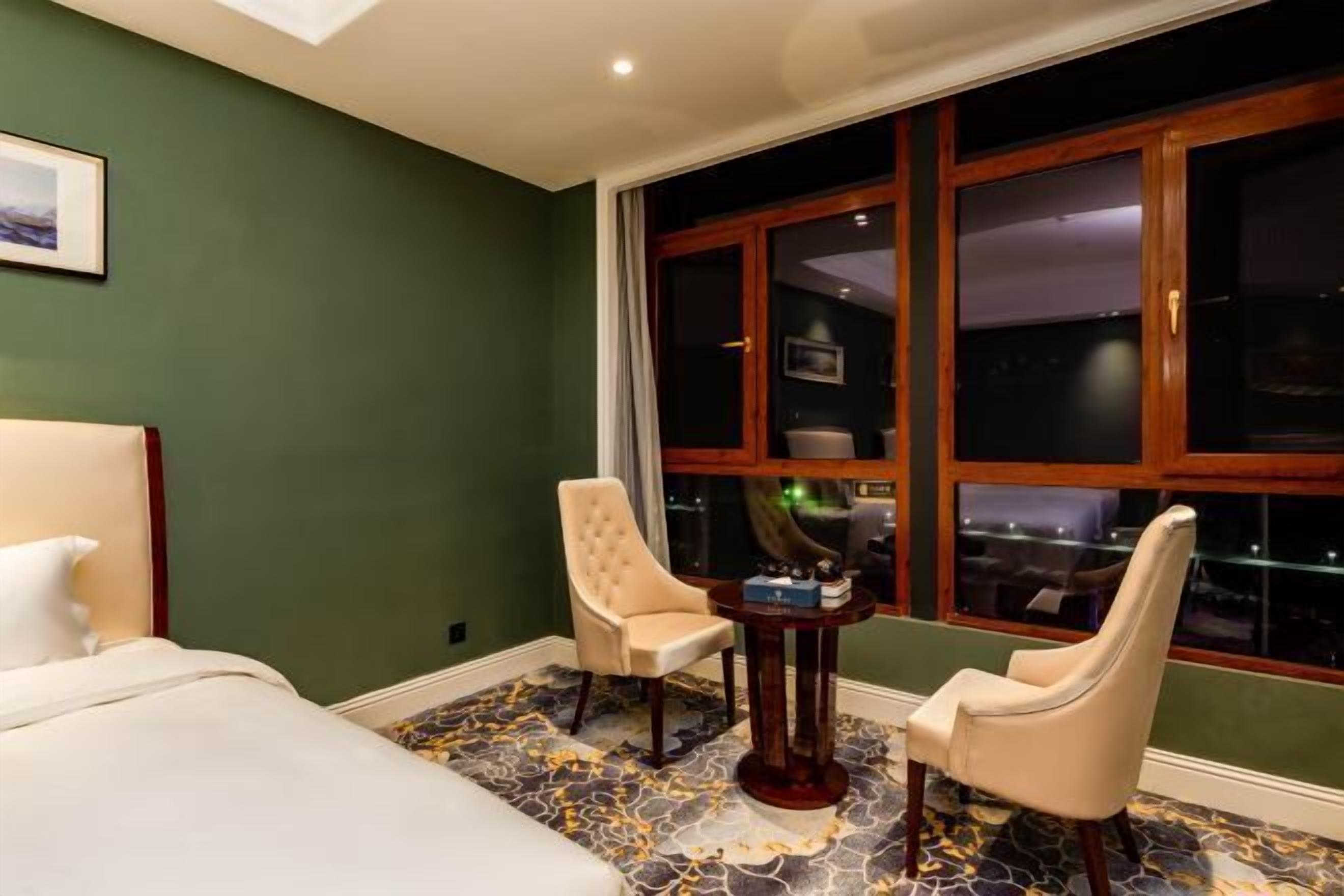 A hotel room with a bed, two chairs, a small table, and a large window with a view of the city at night.