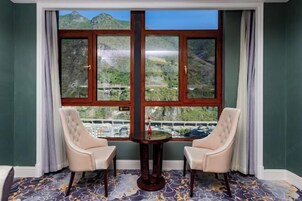 Quarto com uma grande janela que oferece vista para as montanhas, duas poltronas estofadas e uma pequena mesa com um objeto decorativo.