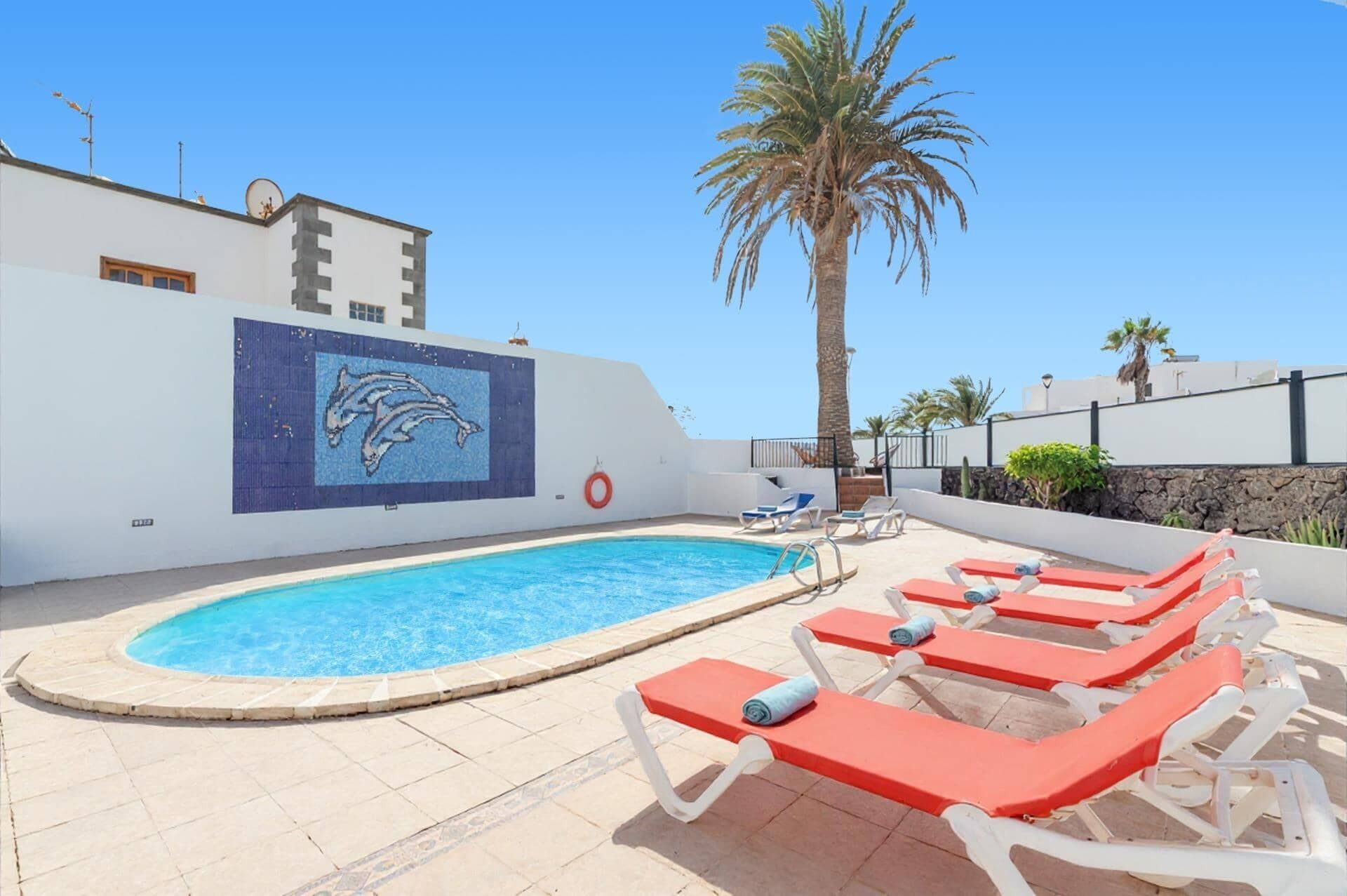 A swimming pool with a dolphin mural, red lounge chairs, and palm trees.