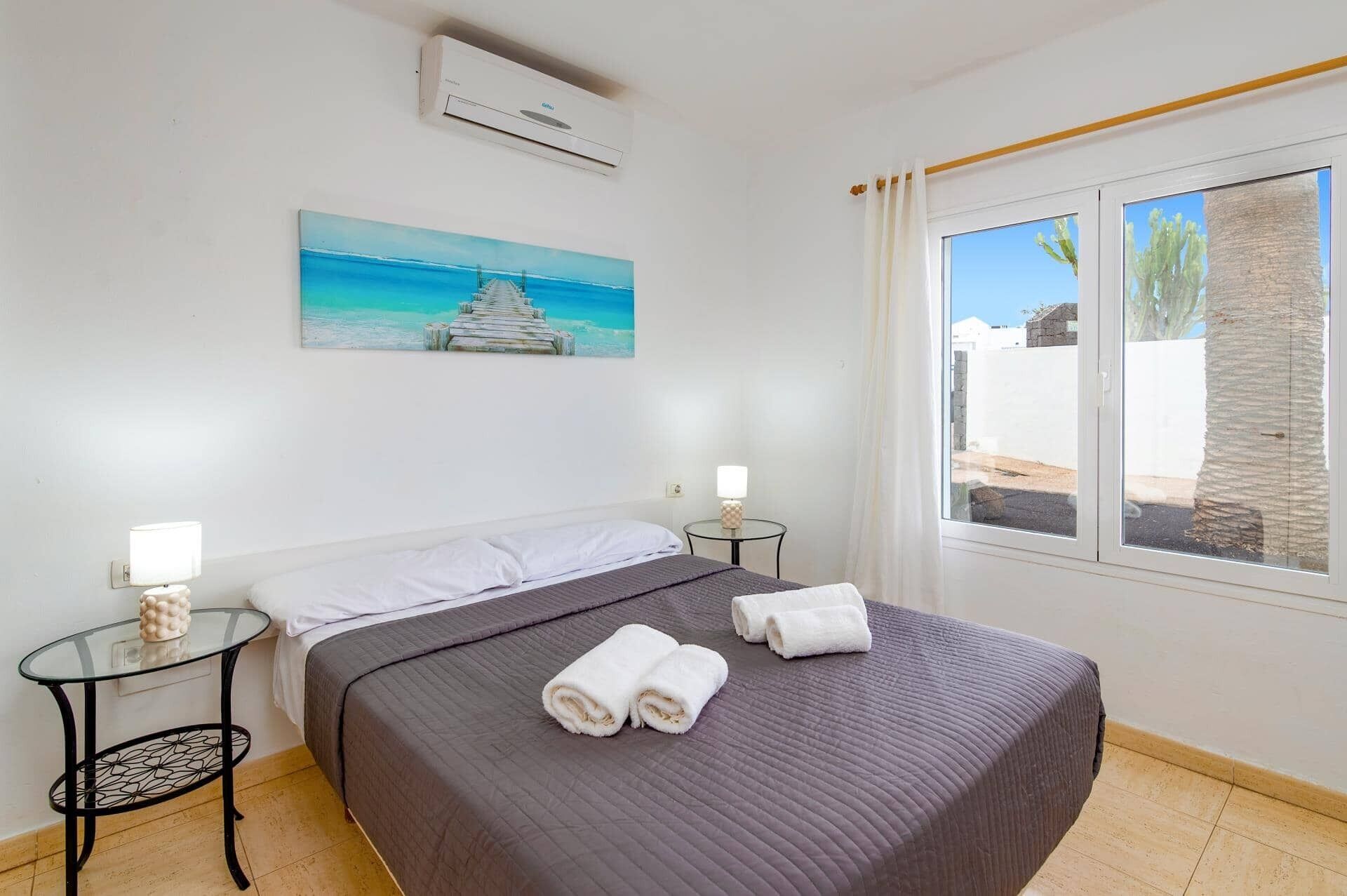 Hotelkamer met een bed, een klein tafeltje met een lamp, een schilderij van een strand en een raam met uitzicht op palmbomen.