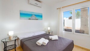 A hotel room with a bed, a small table with a lamp, a painting of a beach, and a window with a view of palm trees.