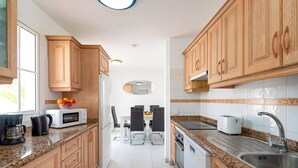 Una cocina moderna con armarios de madera, encimeras de granito, una zona de comedor con mesa y sillas y un refrigerador visible.