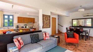 A modern living room with a grey sofa, red armchair, and a dining area with a table and chairs. A kitchen with wooden cabinets and a large window is visible in the background. - Villa Aqua Maris by Villa Plus (Rhodes)
