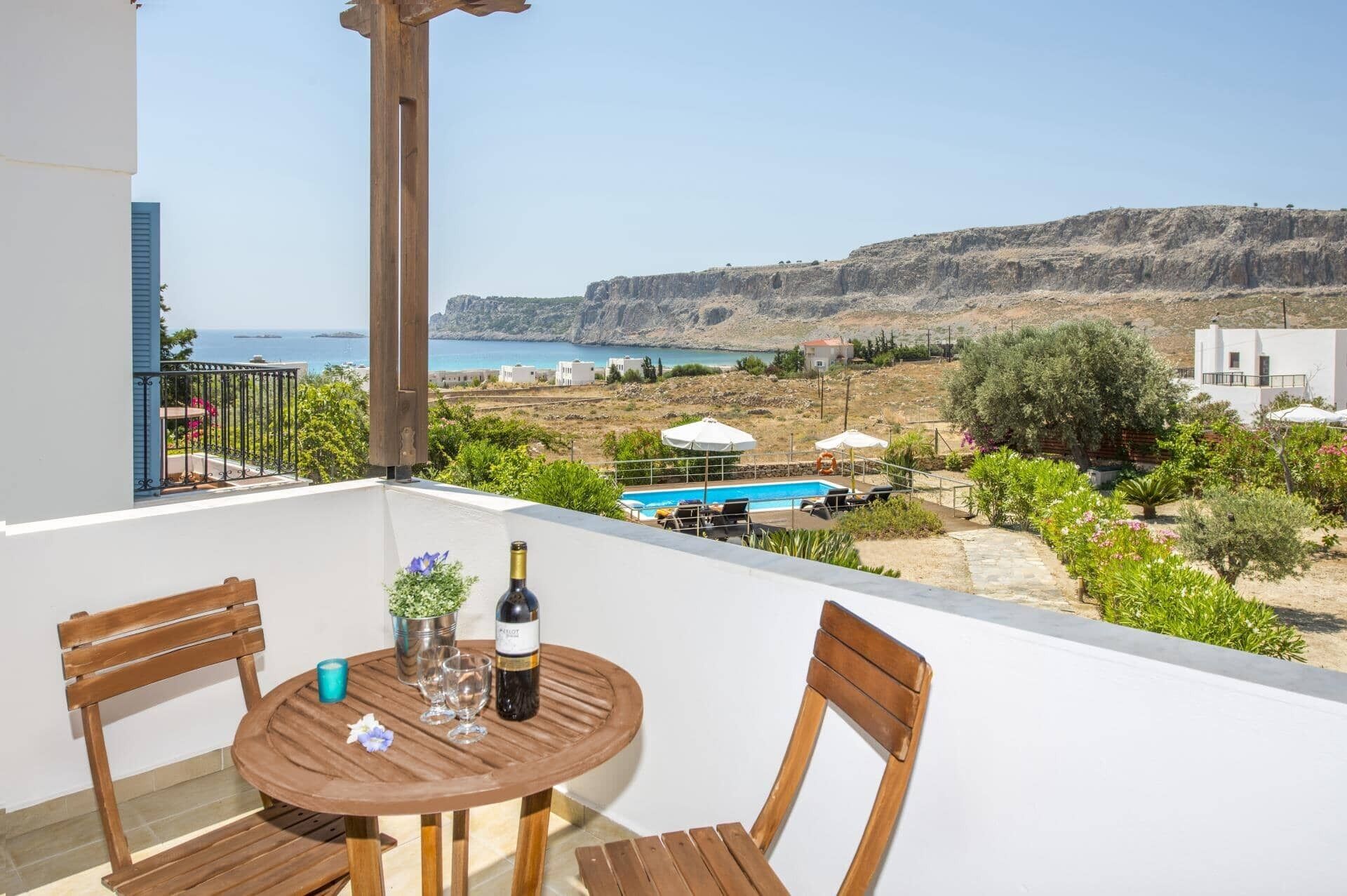 A balcony with a table set for two, overlooking a scenic view of the sea and a mountainous landscape.