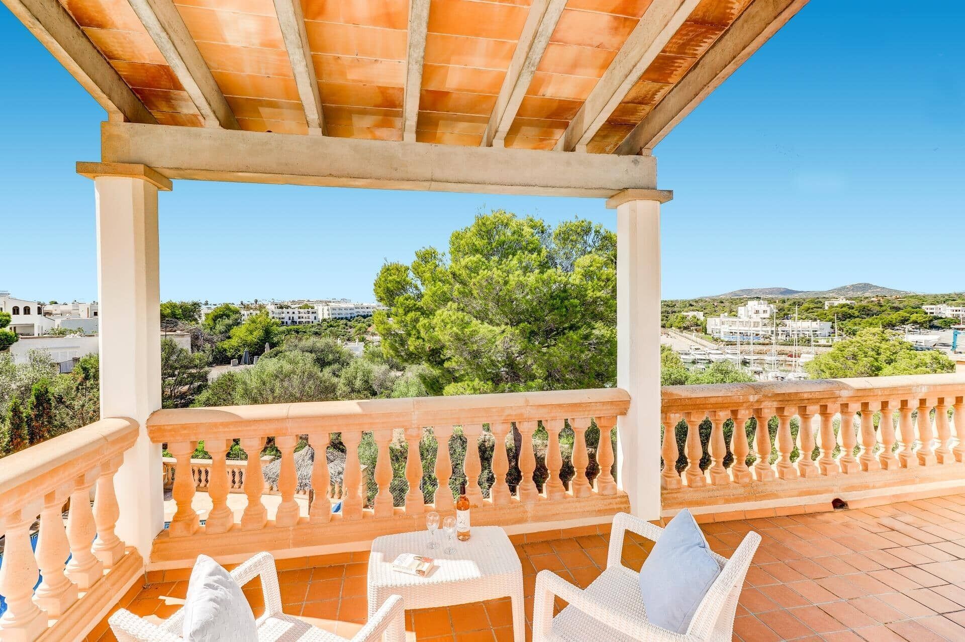 Una terraza con vista a un pueblo costero, que incluye una pérgola, muebles de mimbre blanco y un piso de baldosas.