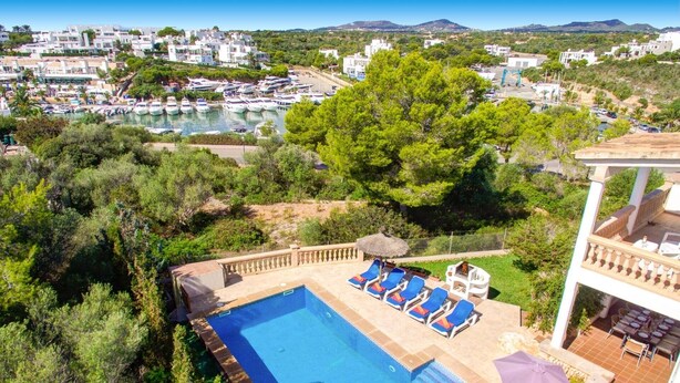A pool area with blue water, surrounded by lounge chairs and umbrellas, overlooking a marina with boats and a coastal town in the background. - Villa Badia D'Or by Villa Plus (Cala d'Or)
