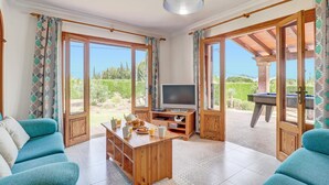 A spacious living room with a television, wooden coffee table, and two teal sofas. Large windows offer views of greenery and a pool. - Villa Bauza by Villa Plus (Mallorca)