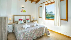 Un dormitorio con una cama grande, dos mesitas de noche, un buró, una ventana con cortinas y vistas a zonas verdes.