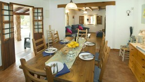 Een eetruimte met een houten tafel voor zes personen, omringd door houten stoelen. Op de achtergrond is een stenen zuil en een houten deur met ruitpanelen zichtbaar.