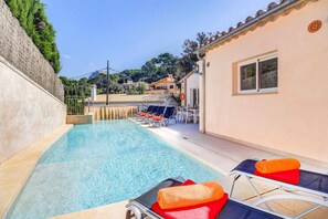 A swimming pool with lounge chairs and a table, surrounded by a building with a tiled roof and a view of the hills.