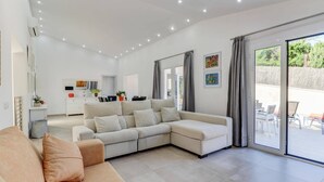 A modern living room with a sectional sofa, a dining area, and a view through large sliding glass doors. - Villa Zaragoza by Villa Plus (Cala Sant Vicenç)