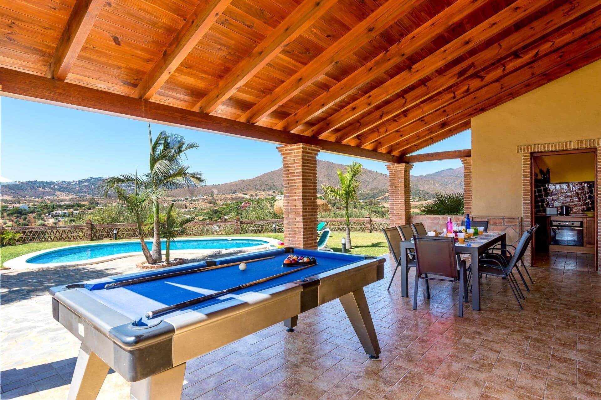 A pool table under a wooden roof with a view of a swimming pool and mountains in the background.