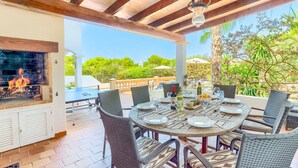 Un patio exterior con una mesa redonda lista para comer, rodeada de sillas de mimbre. Se ve una parrilla empotrada al fondo, y el espacio está cubierto por un techo de madera.