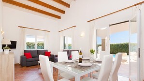 A spacious living area with a glass dining table, white chairs, and a sofa with red cushions. There is a television on a side table and a bottle of wine on the table. - Villa Angelas by Villa Plus (Cap d'Artrutx)