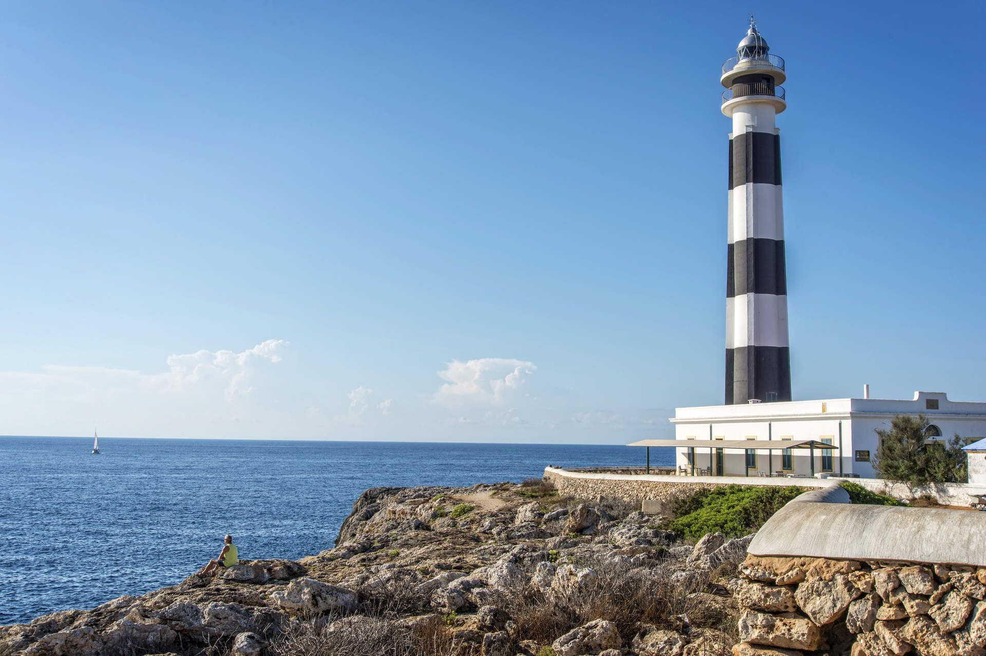 A lighthouse with a white and black striped tower, a stone pathway, and a building with large windows overlooking the sea.