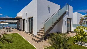 A modern house with a glass staircase, a patio with outdoor furniture, and a well-maintained lawn. - Villa Alana by Villa Plus (Corralejo)