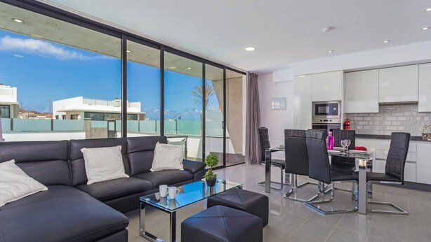 A modern living room with a black leather sofa, a glass coffee table, and a dining area with a black chair and a small table. - Villa Alana by Villa Plus (Corralejo)