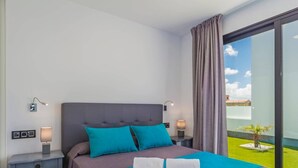 A modern hotel room with a large bed, two pillows, a nightstand, a bedside table, and a view of a green outdoor area through a glass door. - Villa Alana by Villa Plus (Corralejo)