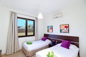 Habitación de hotel con dos camas, una ventana con cortinas, un equipo de aire acondicionado y dos cuadros colgados en la pared.
