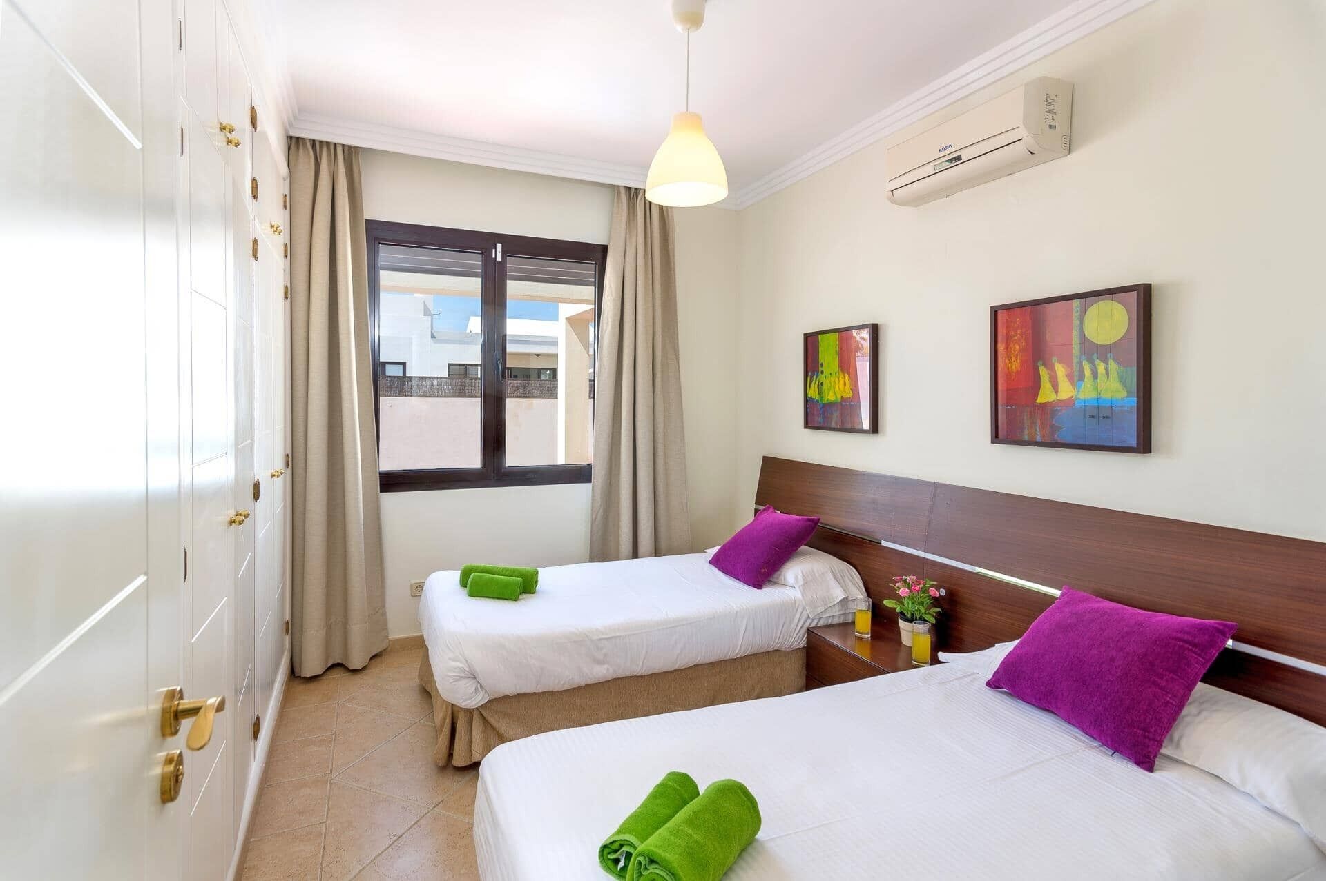Une chambre d’hôtel avec deux lits, une fenêtre avec des rideaux, un appareil de climatisation et deux œuvres d’art accrochées au mur.