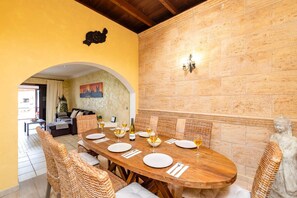 A dining area with a wooden table set for four, surrounded by wicker chairs, a stone wall, and a cityscape painting.