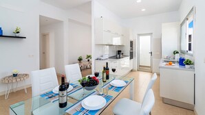 A modern dining area with a glass table set for four, white chairs, and a kitchen in the background. - Villa Yucca Mar by Villa Plus (Playa Blanca)