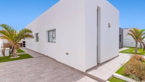 A modern, single-story building with a white exterior and a flat roof. There are two windows visible, one on the left and one on the right side of the building. The building is surrounded by a well-maintained lawn and a few palm trees. - Villa Yucca Mar by Villa Plus (Playa Blanca)