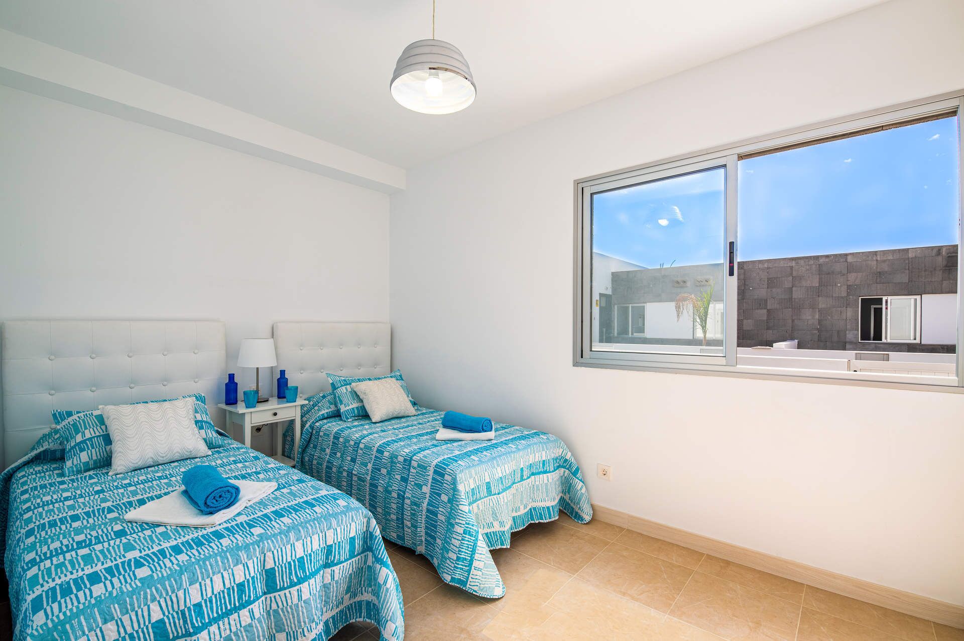 Une chambre avec deux lits, une table de chevet, une lampe et une fenêtre donnant sur des immeubles.