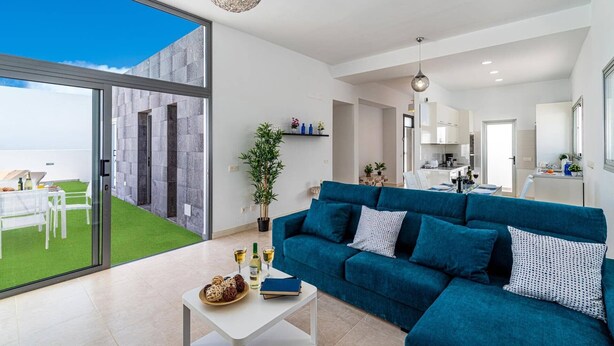 A modern living room with a blue sofa, a dining table with drinks, and a view of a garden through large windows. - Villa Yucca Mar by Villa Plus (Playa Blanca)