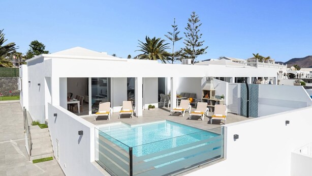 En modern villa med en infinitypool som är omgiven av glas, en utomhusplats och utsikt över palmer och berg.