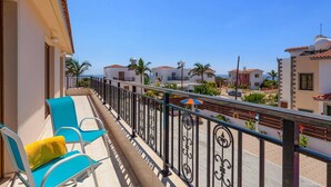 A balcony with blue chairs and a yellow cushion, overlooking a residential area with houses and a clear sky.