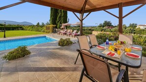 A poolside area with a covered patio, lounge chairs, and a table set for breakfast. - Villa Andriana by Villa Plus (Latchi)