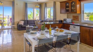 Amplia sala de estar con mesa de comedor para cuatro, cocina con armarios de madera y ventanas grandes que dan a zonas verdes.