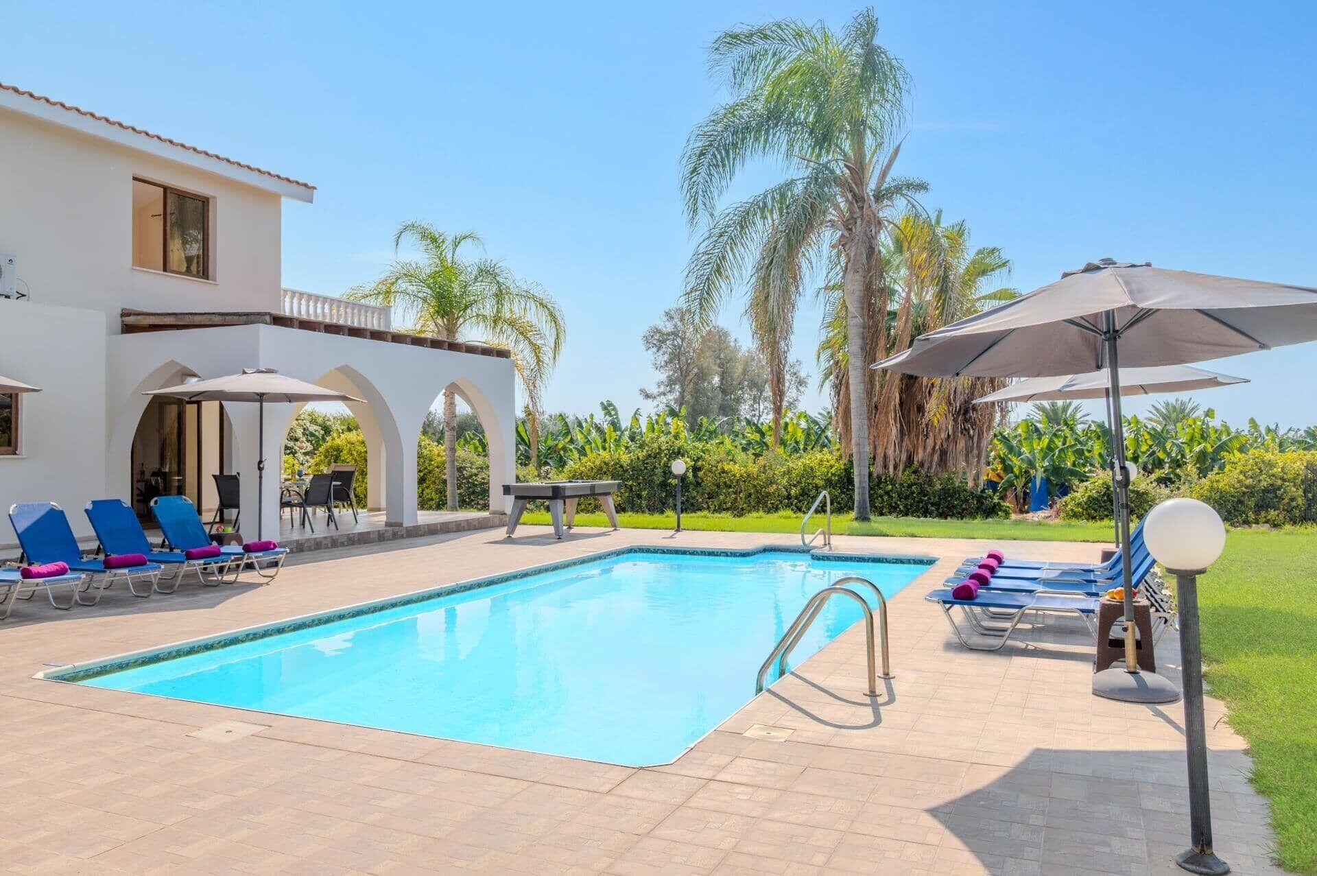 A swimming pool with lounge chairs and umbrellas, surrounded by palm trees and a white building with arches.