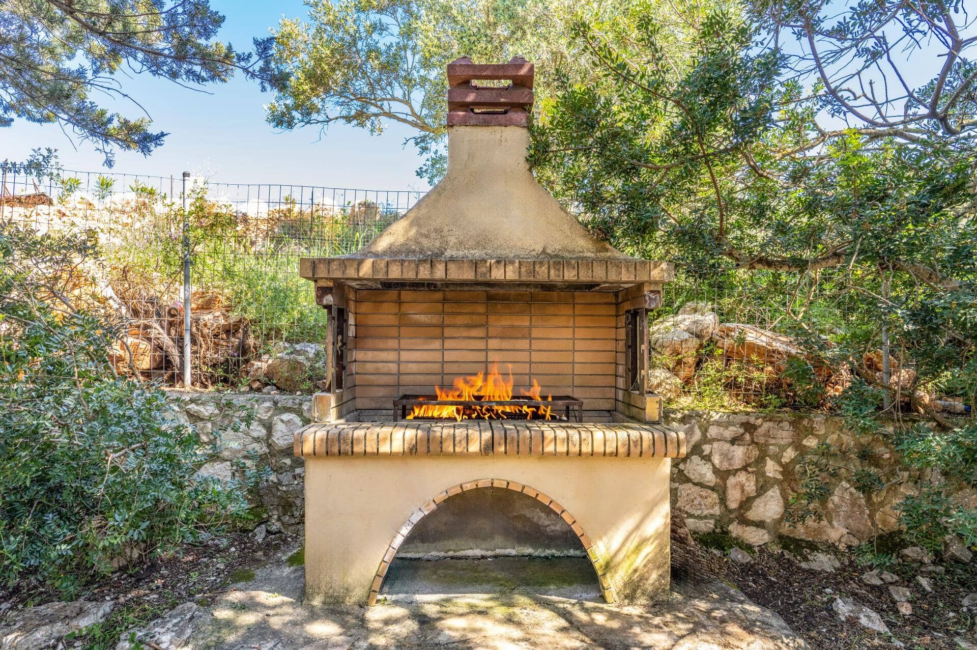 An outdoor stone-built wood-burning stove with a fire burning inside, surrounded by greenery and a stone wall.
