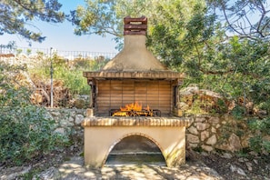 An outdoor stone-built wood-burning stove with a fire burning inside, surrounded by greenery and a stone wall.