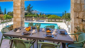 Un espace de restauration en plein air avec une table dressée pour deux personnes, offrant une vue sur une piscine et la mer.