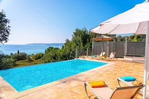 Une piscine avec vue sur la mer, entourée d’une terrasse carrelée agrémentée de chaises longues et de parasols.