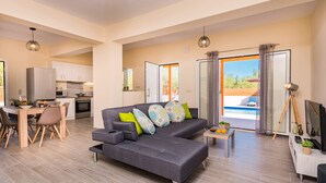 Ein modernes Wohnzimmer mit einer grauen Eckcouch, einem Essbereich mit Holztisch und -stühlen, einer Küche mit weißen Schränken und einer Glasschiebetür zum Poolbereich.