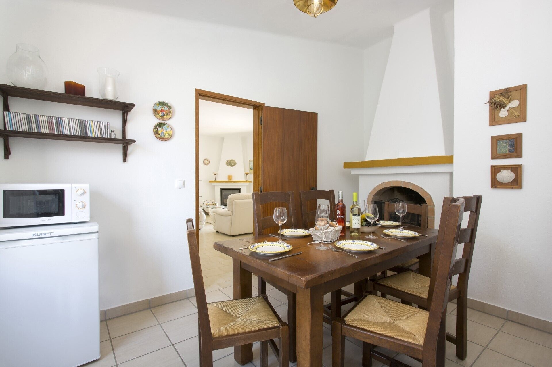 A dining area with a wooden table set for four, surrounded by chairs with woven seats. A white microwave is visible on the left, and a bookshelf with various items is above it.