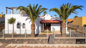 A white building with a red roof, two palm trees, and a stone wall in front.