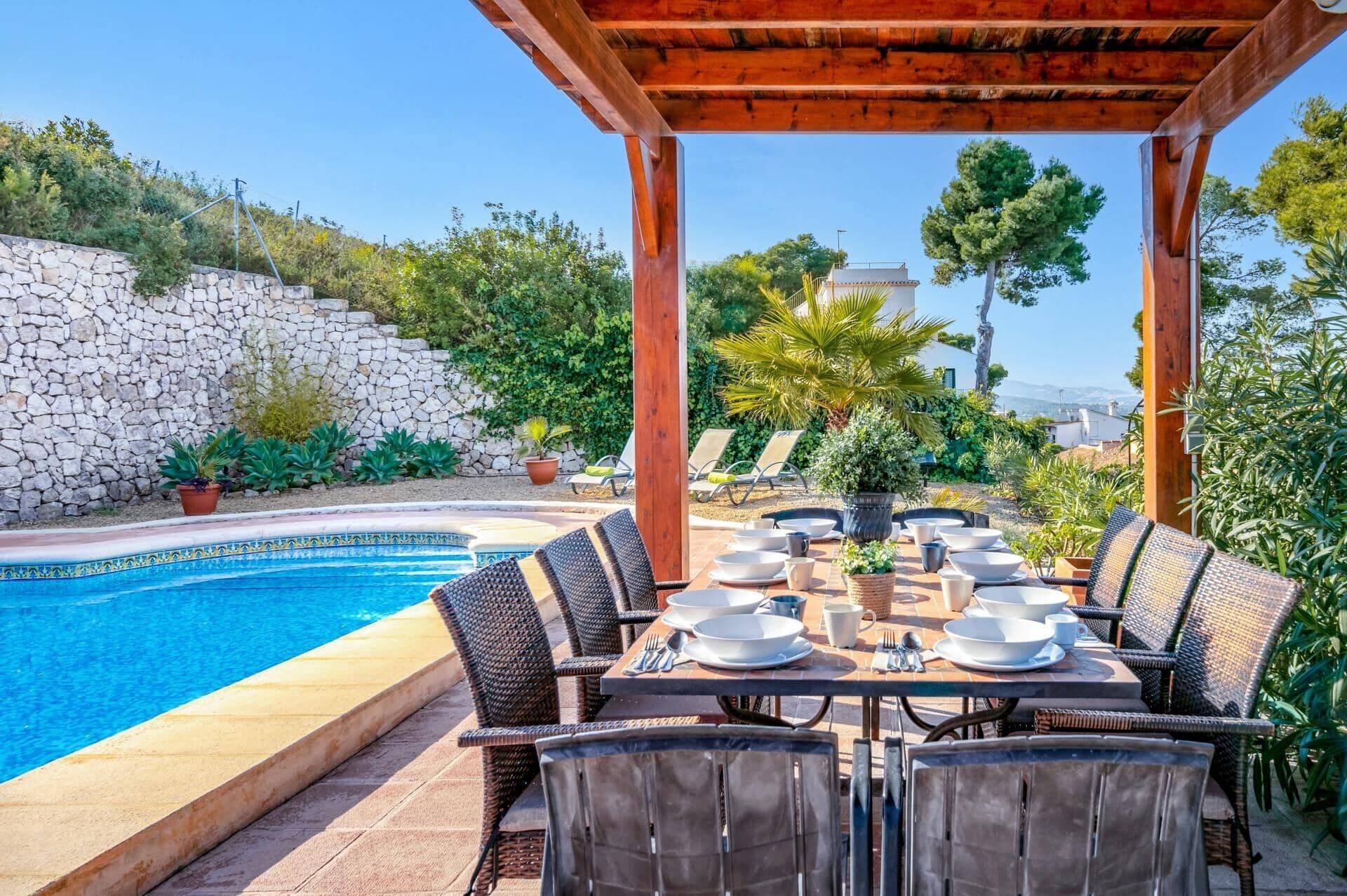 A poolside dining area with a pergola, stone wall, and lush greenery.