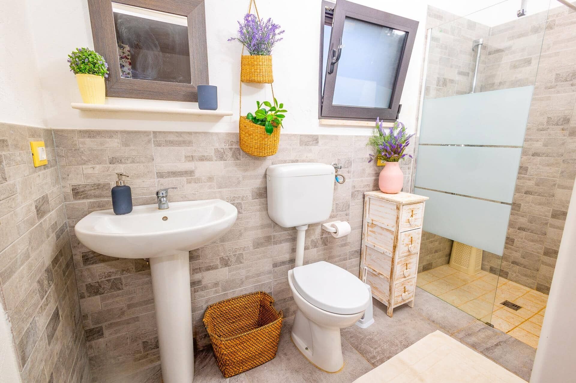 A bathroom with a white sink, toilet, and shower. There are wicker baskets and potted plants on the wall.