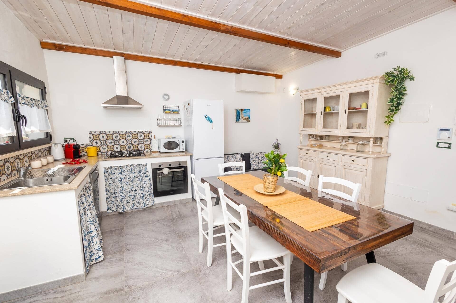 Cozinha compacta com armários brancos, uma mesa de jantar com cadeiras e um frigorífico pequeno.