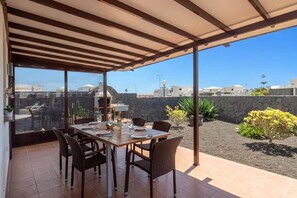 Une terrasse extérieure avec une table dressée pour le repas, des chaises en osier, un barbecue et une vue sur des bâtiments et un mur de pierre.
