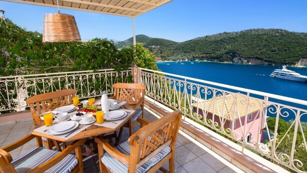 Eine Terrasse mit Frühstückstisch, mit Blick auf eine Küstenbucht mit Booten und einem Kreuzfahrtschiff.