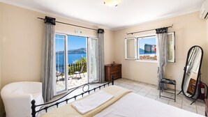 Ein Schlafzimmer mit Bett, Kommode, Stuhl und einem großen Fenster mit Blick aufs Meer.