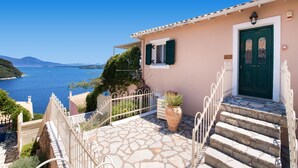 Ein Küstenhaus mit einer Steintreppe, die zu einer grünen Tür führt, einem kleinen Balkon mit Metallgeländer und Blick aufs Meer.