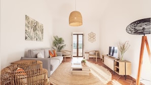 A modern living room with a sofa, coffee table, and wicker chairs. There is a TV, a plant, and a large window. - Quinta da Baleeira (Tavira)
