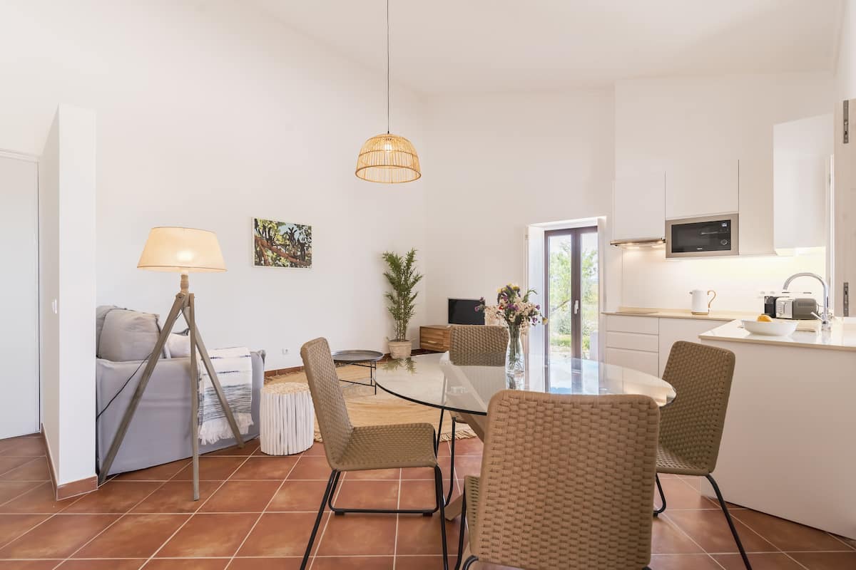 Una cuina moderna amb armaris blancs, microones integrat i zona de menjador amb taula de vidre i cadires de vimet.